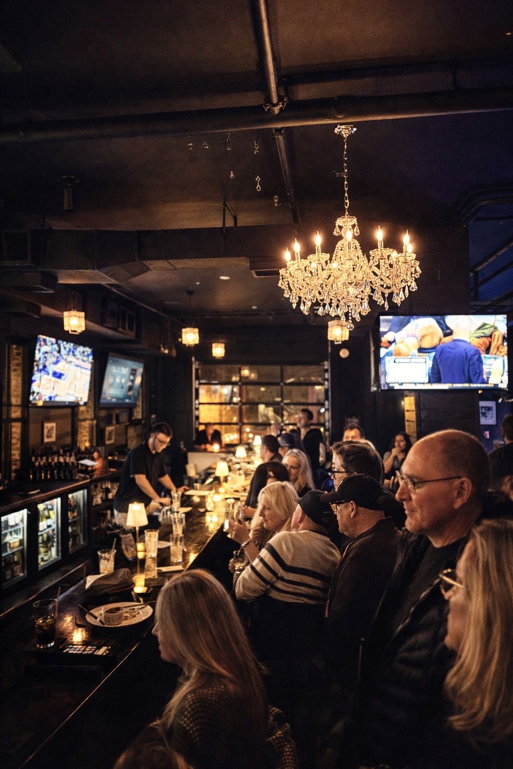 A full bar under the chandelier at Suite 200 in Milton, Georgia — guests along the rail, bartender pouring, games on the screens